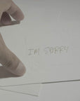 A close-up of a hand presenting a minimalist white "I'm Sorry" note card, highlighting its clean typography and premium paper texture.
