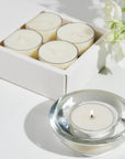 A lit Vaucluse Jasmine tealight candle burning inside a clear glass holder, placed in front of a white product box and delicate white flowers.