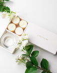 Top-down minimalist flat lay of the open jasmine tealight candle set styled beautifully with fresh white jasmine flowers and green leaves on a white surface.