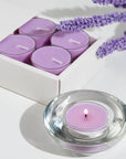A lit purple Vaucluse Lavender tealight candle burning in a clear glass holder, styled with a white product box and a sprig of dried lavender flowers.