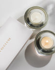 An overhead top-down view of a lit vanilla scented tealight candle softly burning in a clear glass holder alongside a white Vaucluse luxury gift box.