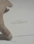 A close-up view of a person's hand interacting with the elegant white Let's Celebrate greeting card against a clean, minimalist background.