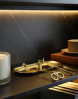 An elegant gold candle care set elegantly arranged on its matching tray resting on a dark modern bookshelf next to a ribbed candle.