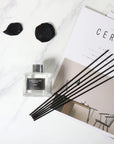 An overhead flat lay view of a clear glass Vaucluse reed diffuser bottle with a cluster of black reeds arranged on a stylized magazine with dark rose petals on a marble counter.
