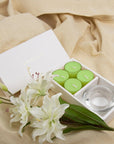 Elegant lifestyle layout showing the green lily tealight set and clear glass holder nestled on a soft, draped beige linen fabric with fresh white lilies.