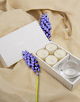 Elegant aromatherapy lifestyle scene featuring the white musk tealight gift box styled on draped beige linen fabric with purple floral accents.
