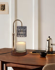 A lit smoky grey crystal candle warmer lamp casting ambient light over a jar candle on a dark wooden table next to a silver coffee moka pot.