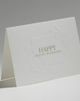 An elegant, freestanding white housewarming greeting card showcasing a detailed blind-embossed floral frame around the central gold typography.