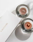 Overhead view of two brown Vaucluse Cinnamon tealight candles in circular glass holders, with one lit and casting a warm glow next to white brand packaging.