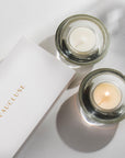Top down view of two Vaucluse Jasmine scented tealight candles in circular glass holders, one lit with a warm flame, next to a minimalist white brand package.