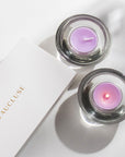 Overhead top down view of two purple Vaucluse Lavender tealight candles in circular glass holders, with one lit and glowing next to a minimalist white brand package.