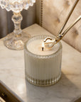 Trimming a candle wick with a polished silver wick trimmer tool on a lit white candle in a ribbed glass jar.