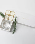Minimalist top-down view of the open vanilla tealight set and glass candle holder, beautifully styled with fresh rosemary sprigs on a crisp white surface.