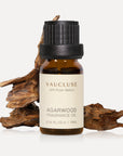 A 10ml amber glass bottle of Vaucluse Agarwood Essential Oil positioned in front of natural textured pieces of raw oud wood.