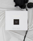 Top down view of the white Vaucluse Black Rose luxury candle gift box resting on soft draped fabric next to a dark black rose stem.