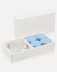 An open luxury white gift box displaying a set of four light blue breeze tealight candles next to a round clear glass candle holder.