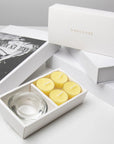 Premium retail unboxing display featuring the open yellow lemon tealight set resting against stacked white gift boxes with gold text.