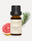 A 10ml amber glass bottle of Vaucluse Grapefruit Cedar Essential Oil backed by a fresh pink grapefruit slice and a green pine needle sprig.