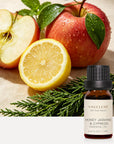Vaucluse Honey Jasmine and Cypress essential oil bottle styled alongside sliced red apples, lemon halves, and green cypress branches.