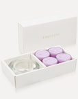 An open luxury white gift box elegantly presenting a set of four light purple lavender tealight candles alongside a thick clear glass candle holder.