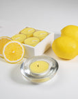 A luxury white Vaucluse 16-pack box sliding open to reveal a tray of yellow lemon tealight candles, shown next to a burning candle in a clear glass holder and whole fresh lemons.