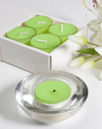 A light green lily scented tealight candle in a clear glass holder shown in the foreground with a partial view of the sliding box tray and white lilies.