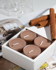 A small white box revealing four brown Vaucluse Cinnamon spiced tealights, arranged on an open magazine with decorative cinnamon sticks.