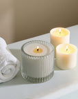A ribbed glass Vaucluse candle alongside two smaller lit votive candles and a white rolled towel on a clean spa ledge.