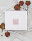 Top down view of the white square Vaucluse Lychee candle packaging box with a pink label, sitting on soft draped fabric next to fresh lychees on stems.
