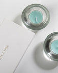 An overhead top-down view of teal ocean scented tealight candles resting in elegant clear glass holders next to a gold-embossed white Vaucluse gift box.
