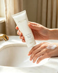 Woman holding Vaucluse Rose Velvet hand cream tube after applying moisturizing cream on her skin by a marble bathroom sink.