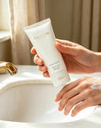 Woman holding Vaucluse Rose Velvet hand cream tube after applying moisturizing cream on her skin by a marble bathroom sink.