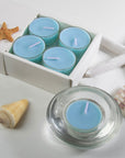 A light blue sea breeze scented tealight candle in a clear glass holder in the foreground, styled with coastal seashells and a starfish next to the tray packaging.