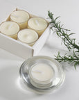 A luxury white Vaucluse 16-pack box sliding open to reveal a tray of clear cup vanilla tealight candles, shown next to a burning candle in a clear glass holder and a fresh sprig of rosemary.