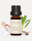 A 10ml amber glass bottle of Vaucluse Wood Sage Sea Salt Essential Oil backed by green sage leaves and a wooden scoop filled with coarse sea salt.