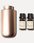 A minimalist rose gold waterless nebulizing aroma diffuser displayed next to two 10ml amber bottles of English pear and freesia essential oil.
