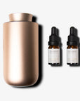 A premium rose gold waterless diffuser shown next to two small amber glass essential oil dropper bottles with clean cream color labels.