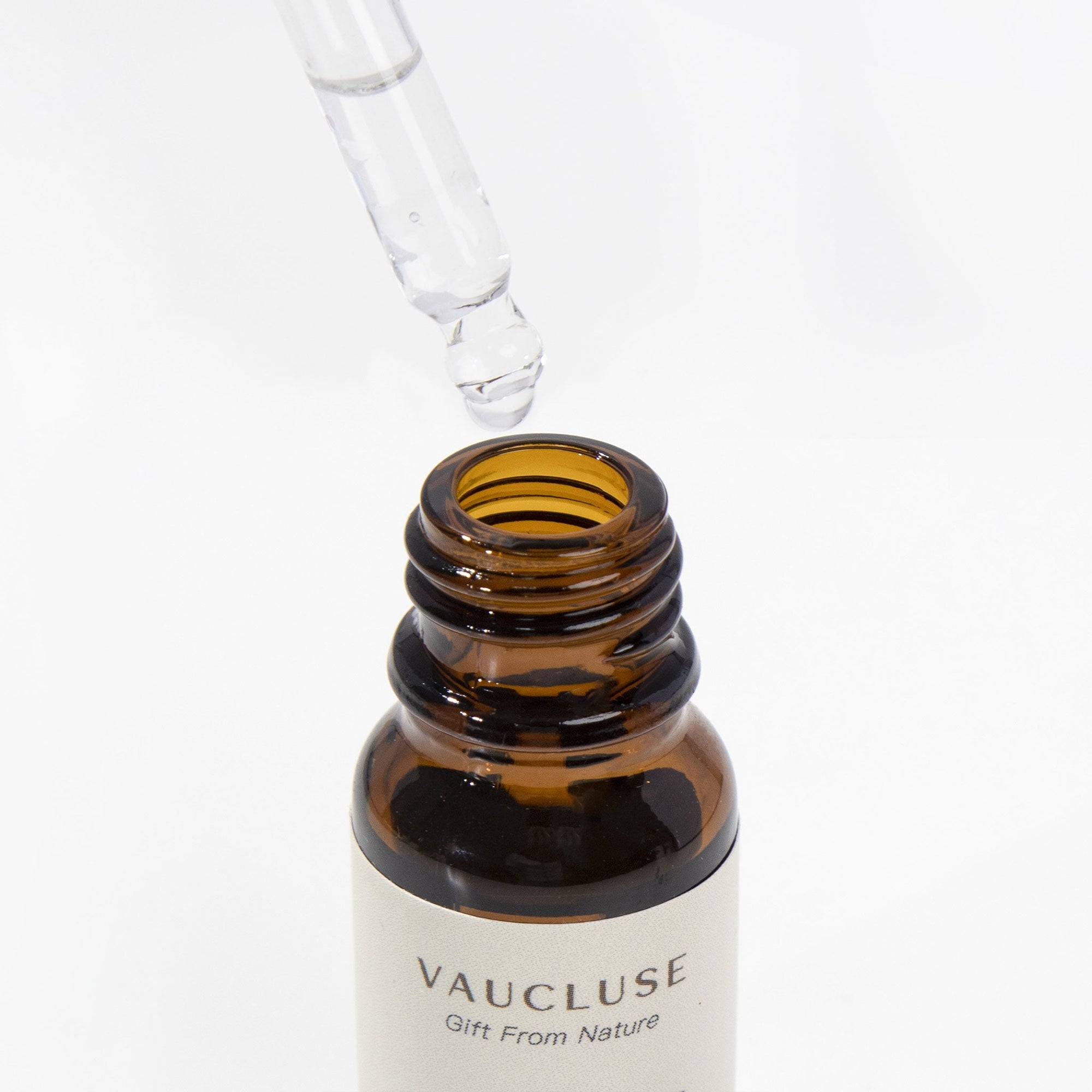 A glass pipette dropper hovering above an open amber glass bottle of Vaucluse essential oil against a crisp white background.