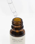 A clear glass pipette dropper dispensing a clean liquid drop into an open amber glass bottle of Vaucluse bergamot essential oil.