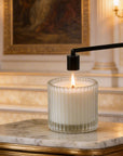 A matte black bell candle snuffer being used to safely extinguish the flame of a luxury white candle on a marble table.