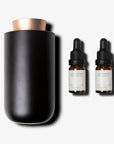 A matte black waterless aromatherapy diffuser with a rose gold accent lid standing next to two amber glass essential oil dropper bottles.