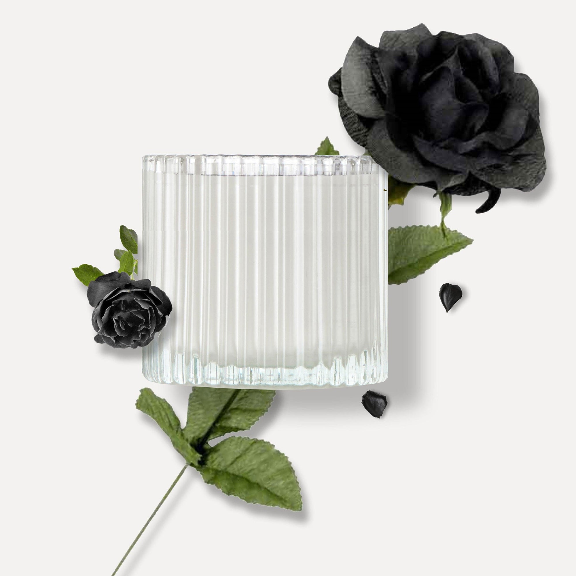 Top view of a Vaucluse ribbed glass candle jar surrounded by dark green foliage and black roses, highlighting the product's primary fragrance notes.