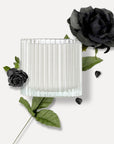Top view of a Vaucluse ribbed glass candle jar surrounded by dark green foliage and black roses, highlighting the product's primary fragrance notes.