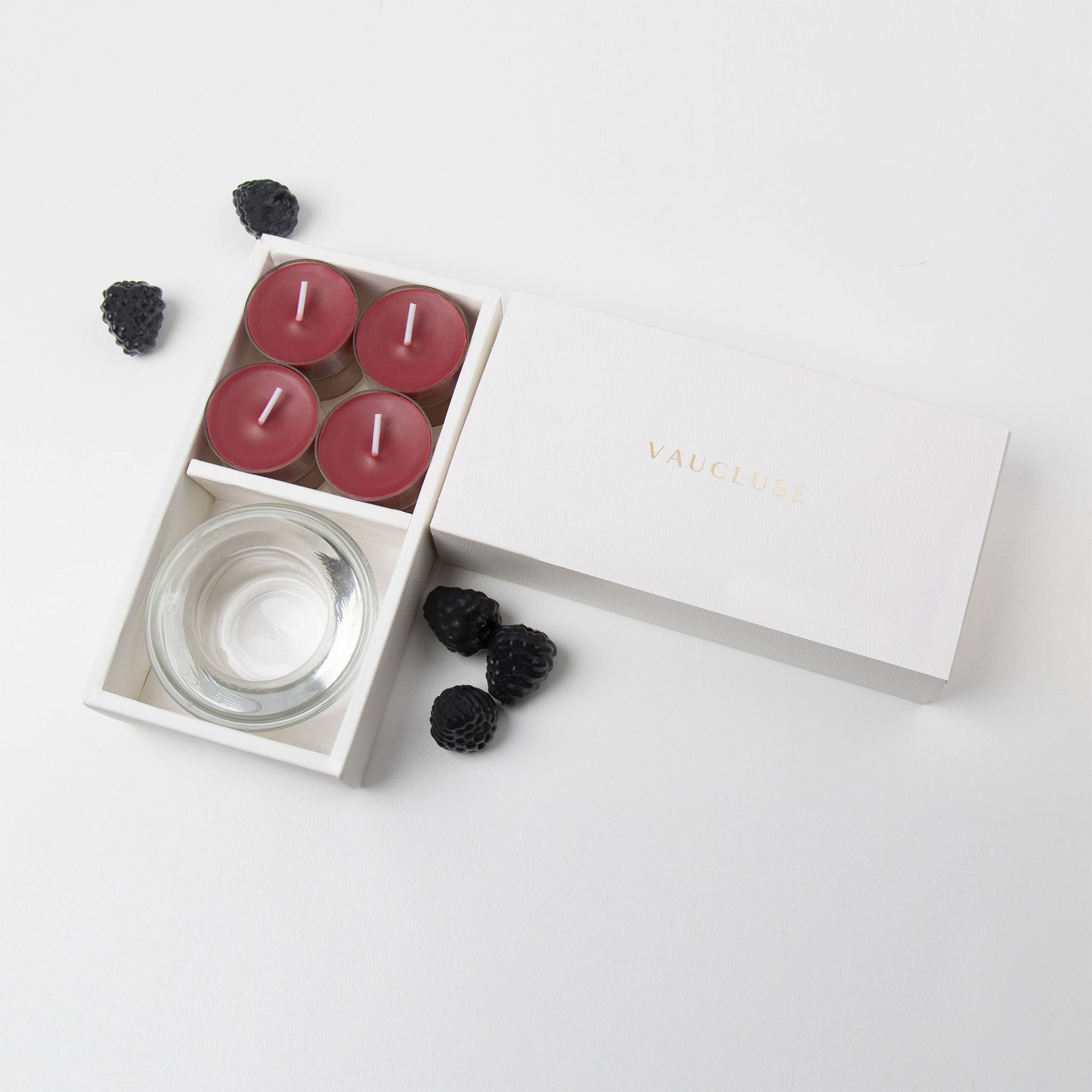 Minimalist overhead flat lay of the open dark red tealight candle set styled with scattered fresh ripe blackberries on a clean white background.