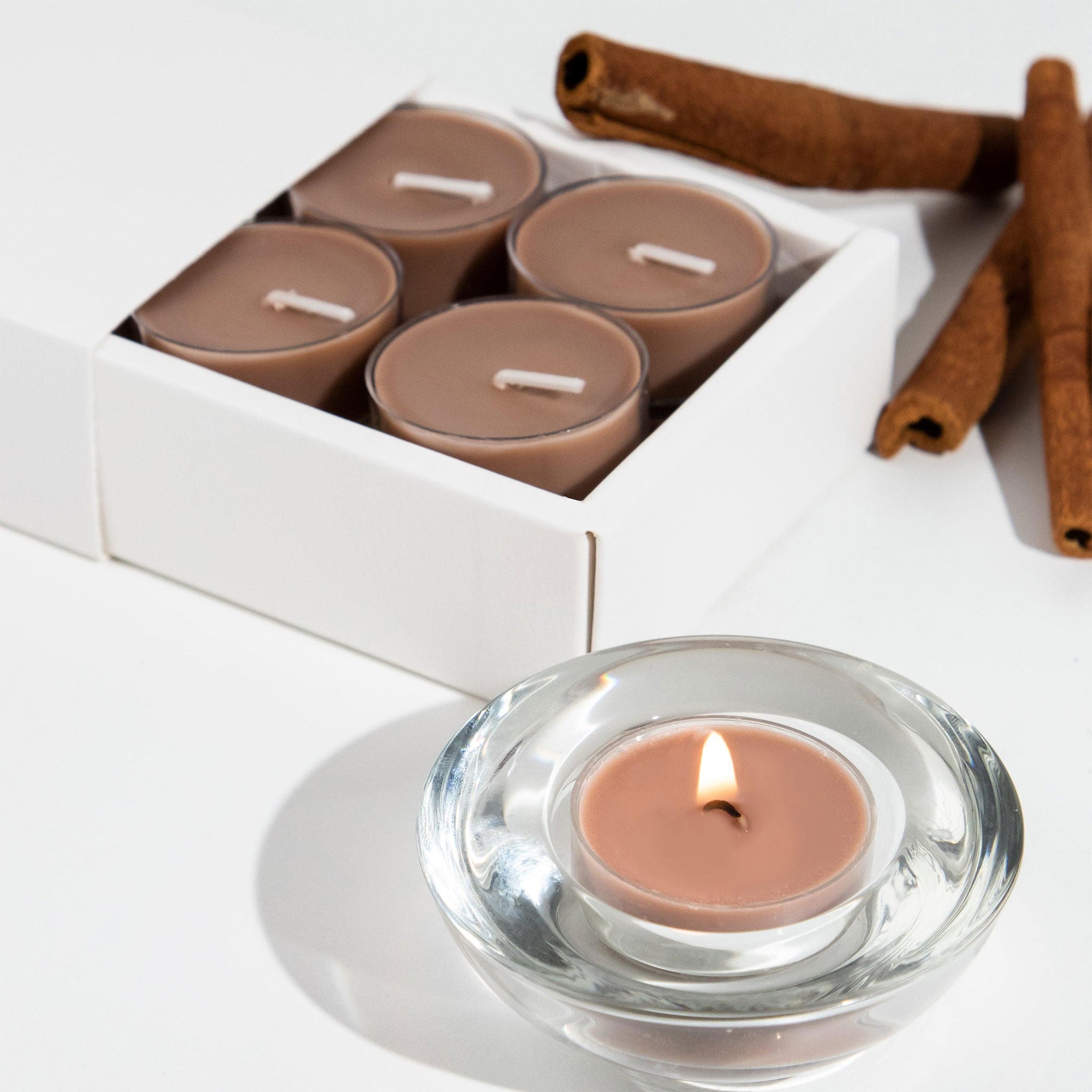 A lit brown Vaucluse Cinnamon tealight candle burning in a clear glass holder, styled next to cinnamon sticks and product packaging.