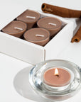 A lit brown Vaucluse Cinnamon tealight candle burning in a clear glass holder, styled next to cinnamon sticks and product packaging.