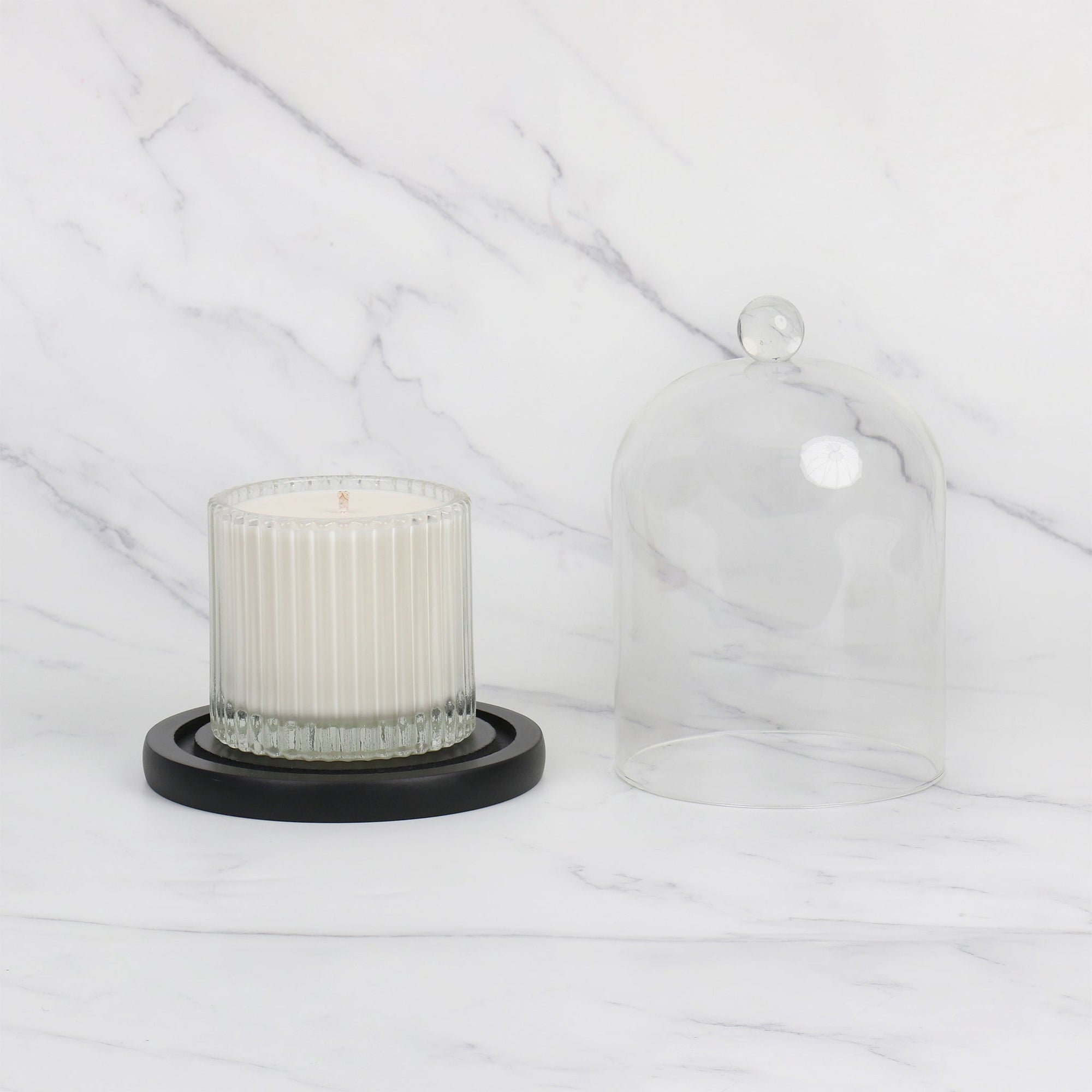 Side-by-side view of a glass cloche lid next to a white ribbed soy candle resting on the round black wooden base over a marble tabletop.