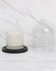 Side-by-side view of a glass cloche lid next to a white ribbed soy candle resting on the round black wooden base over a marble tabletop.