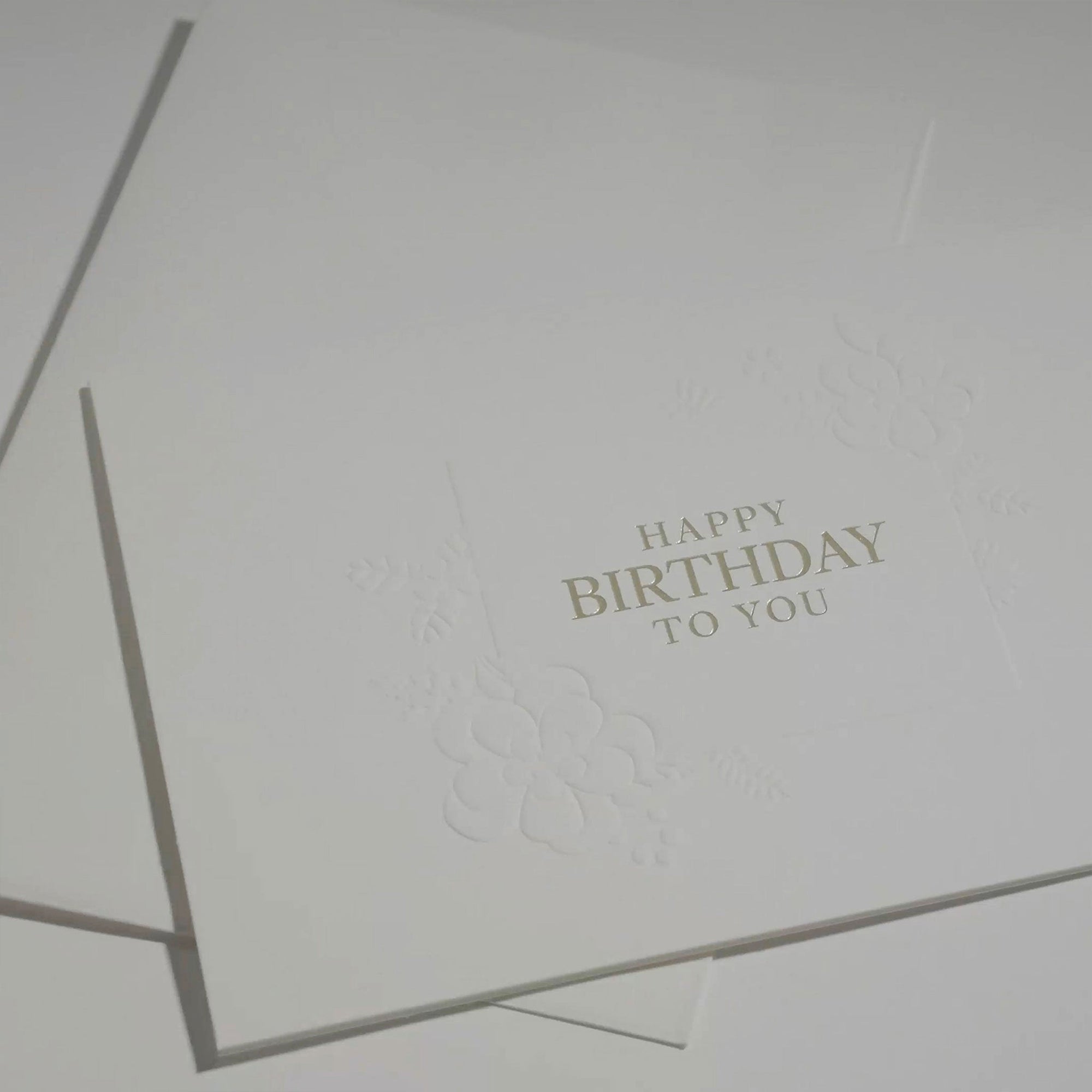 Side angle detail shot focusing on the delicate blind-embossed floral illustrations and fine paper quality of an upscale birthday card.