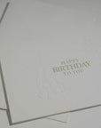 Side angle detail shot focusing on the delicate blind-embossed floral illustrations and fine paper quality of an upscale birthday card.